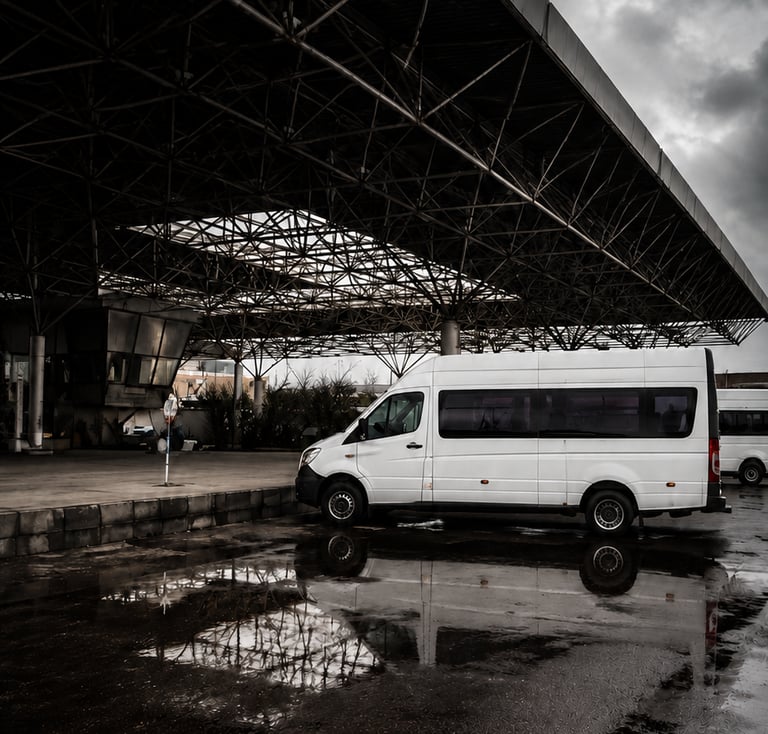 White passenger van parked under a large metal canopy reflected in a rain puddle under a cloudy sky.