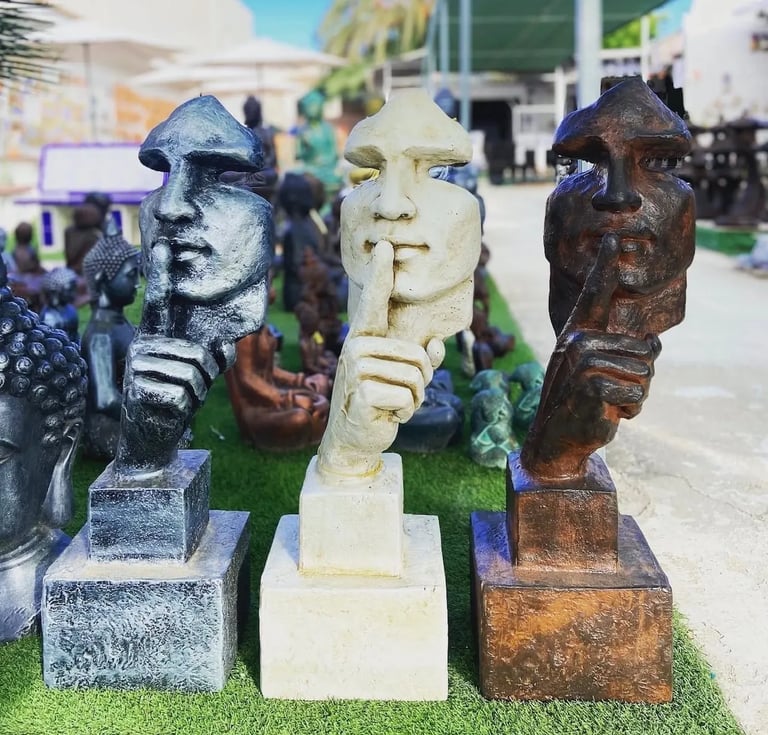 Three silence sculptures in silver, white, and bronze displayed in an outdoor garden decor shop.