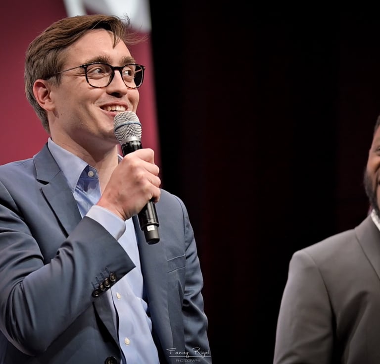 A smiling male speaker in a blue suit holding a microphone at a conference stage.