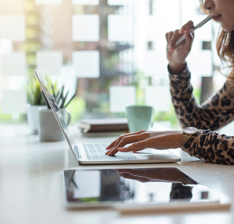 Professional woman holding a pen near her mouth, working on a laptop on a desk with a digital tablet, plant, and coffee cup.