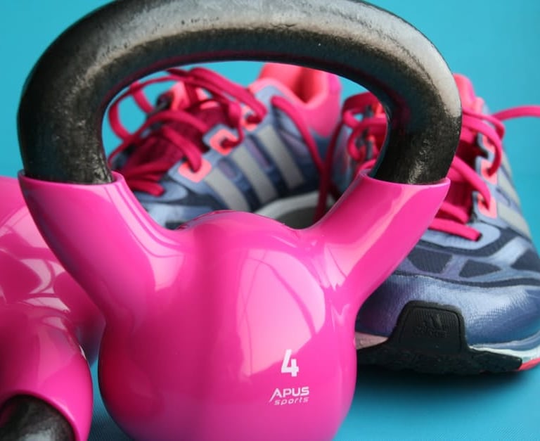 A pink kettlebell sits on a blue surface with tennis shoes in the background.