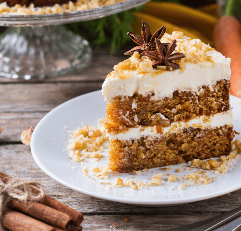Slice of layered carrot cake with cream cheese frosting, chopped nuts, and star anise on a white plate. 