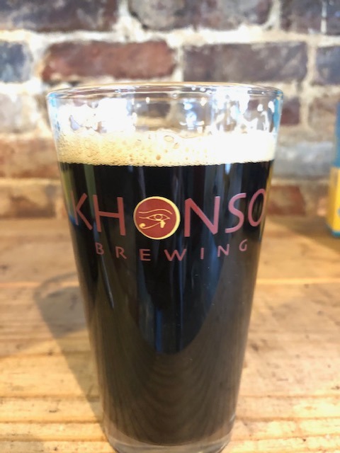 A pint glass of dark craft beer from Khonso Brewing with a frothy head on a rustic wooden table.
