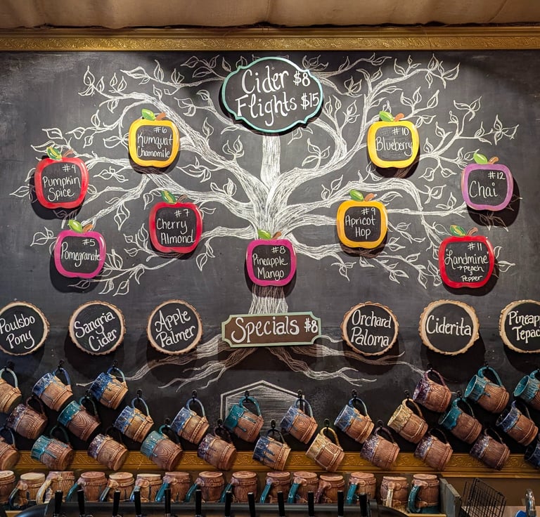 Hand-drawn chalkboard menu featuring hard cider flight flavors and specials above a row of ceramic mugs.