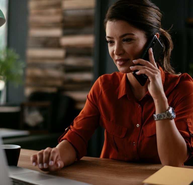 A woman in a red blouse, sitting at her desk talking on the phone.