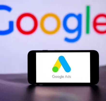 Celular en primer plano mostrando Google Ads, con una computadora desenfocada al fondo con "Google"