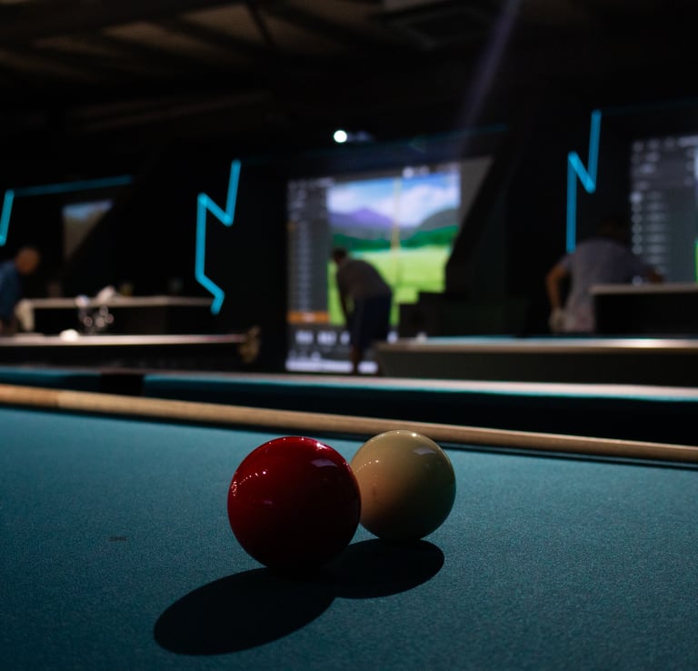 A pool table in the foreground with an indoor golf simulator bay behind it