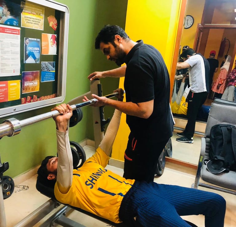 A personal trainer spotting a young man performing a barbell bench press in a gym setting.