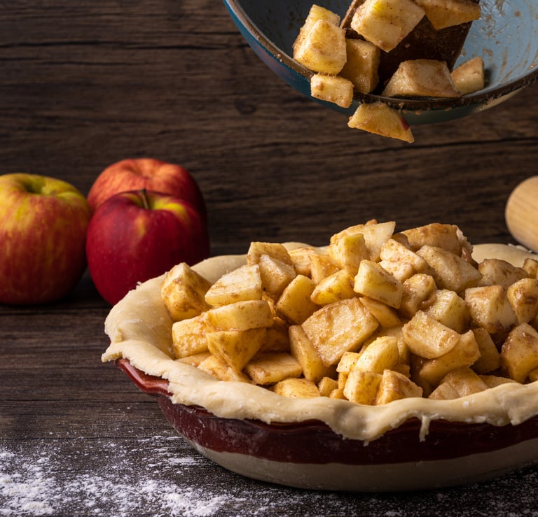 Diced spiced apples being poured from a bowl into an unbaked pie crust next to whole red apples on dark wood. 
