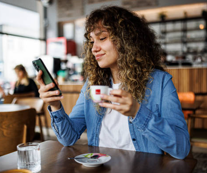 A curly-haired woman in a blue denim shirt using her smartphone while enjoying an espresso at a cafe.
