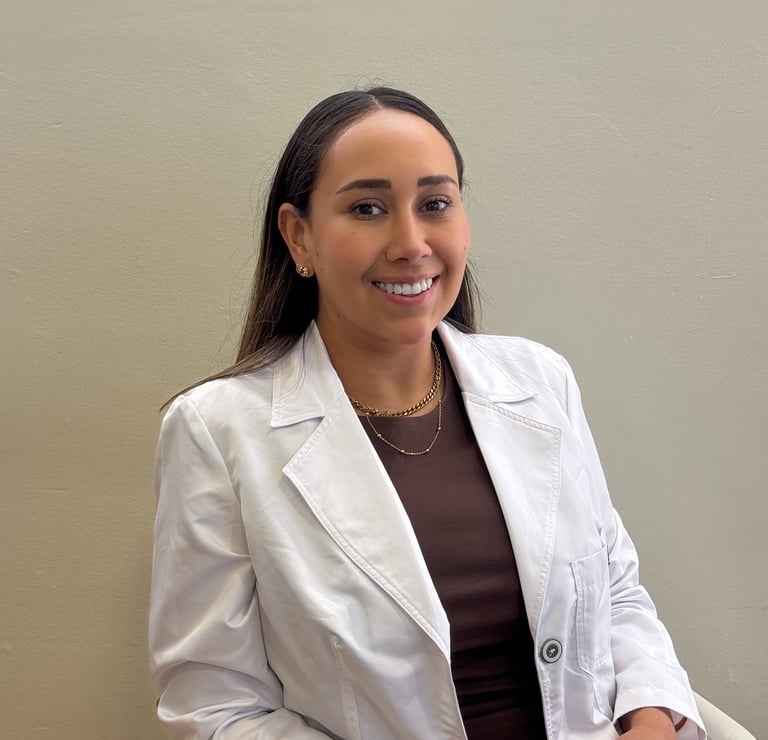 A smiling female medical professional in a white doctor lab coat posing in a clinic office setting.
