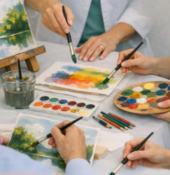 Mãos de pacientes e terapeuta pintam em mesa com tintas e telas em arteterapia hospitalar