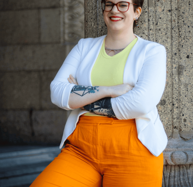 ein Foto von Swana Schuchmann. Sie trägt einen weißen Blazer und lehnt sich an eine Säule, lachend.