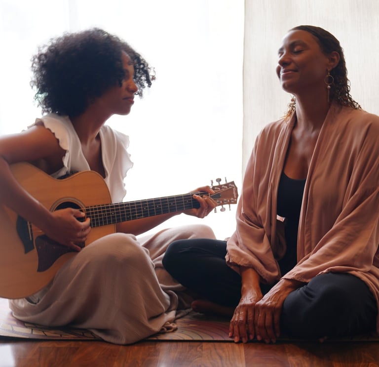 two women in front of a bright window sitting on a carpet playing a guitar.