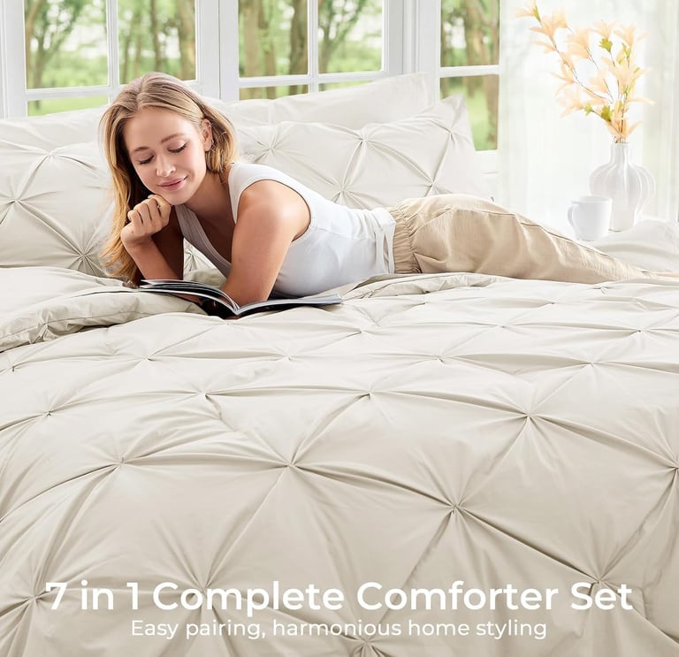 A woman relaxes on a cream 7-piece pinch pleat comforter set while reading a book in a sunlit bedroom.