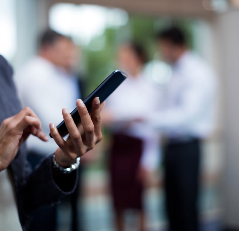 Person using a smartphone with colleagues conversing in the background.