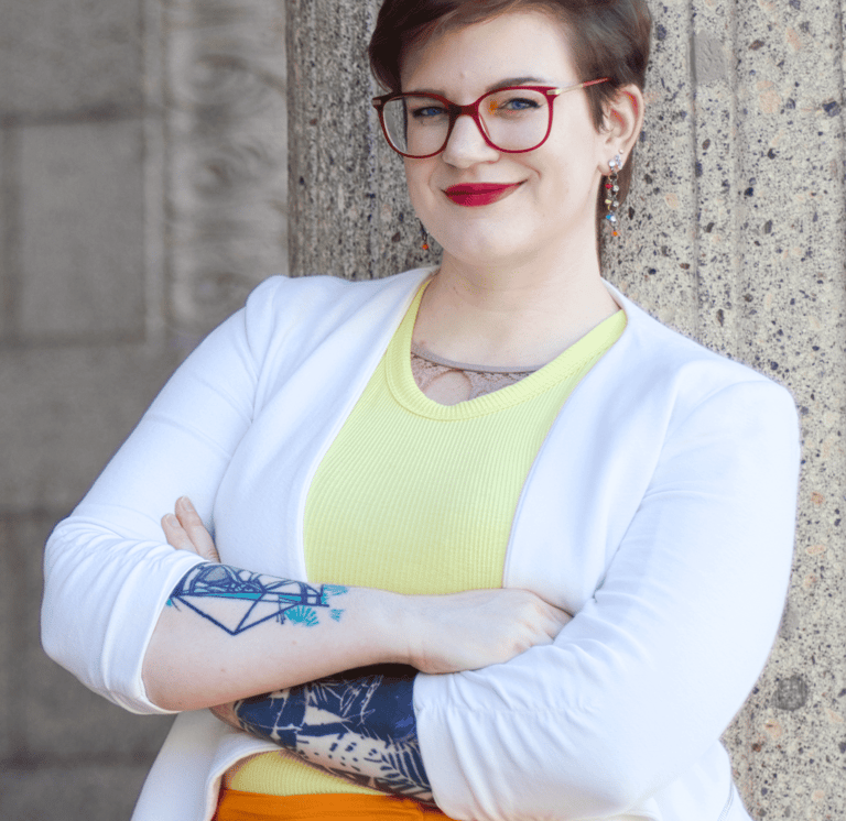 Swana Schuchmann mit Tattoos auf ihren Armen, roter Brille, gelben Top und weißem Blazer, lächelnd