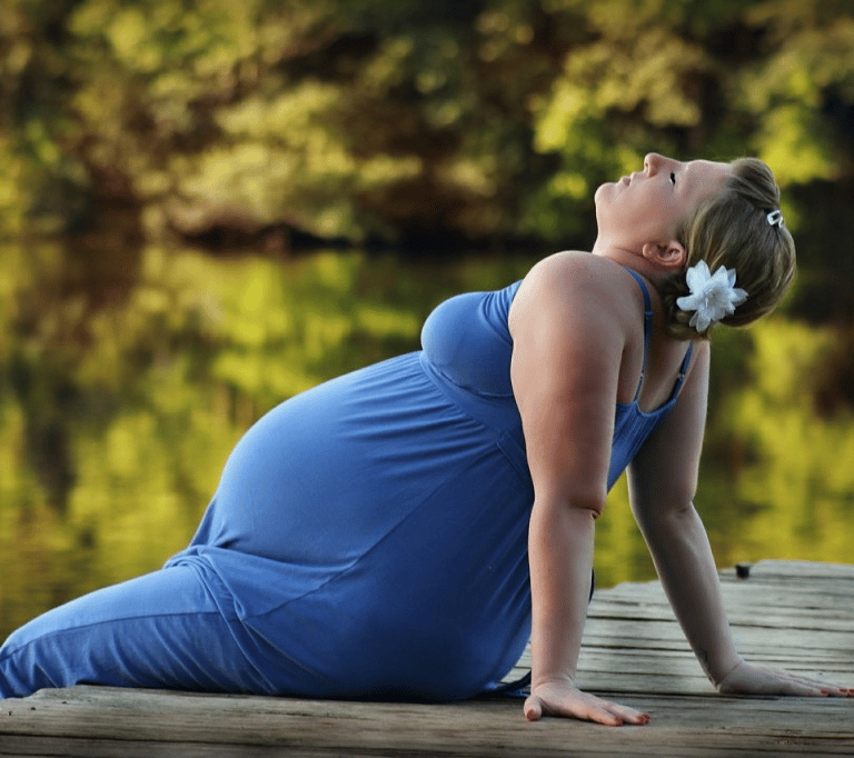 A pregnant woman in a blue dress sits on a wooden dock by a peaceful lake at sunset.