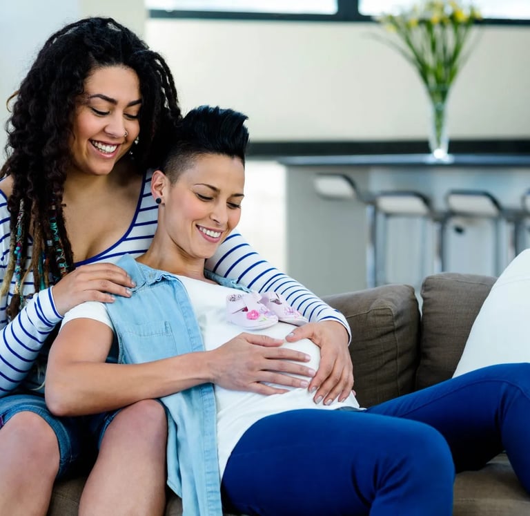 A happy lesbian couple sitting on a sofa together while touching a pregnant belly with baby shoes.