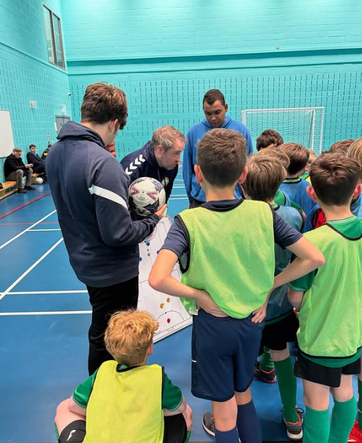 Indoor coaching session in a sports hall with a group of young footballers gathered around coaches d