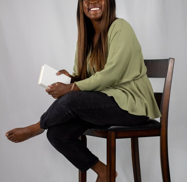 Esther Osaheni, founder of Incurable Optimist, seated on a wooden chair holding a book and smiling