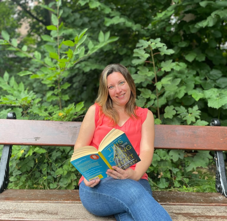 Portrait photo of Jen Ruthe sitting on a park bench in a red top, holding a book.