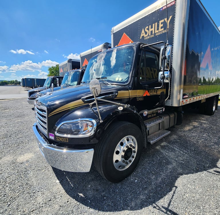 Ashley Furniture delivery trucks parked in a gravel lot on a sunny day.