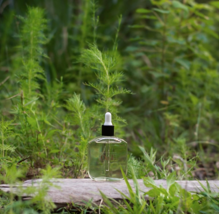 A clear glass dropper bottle of facial oil sitting on a piece of wood in a lush green garden.
