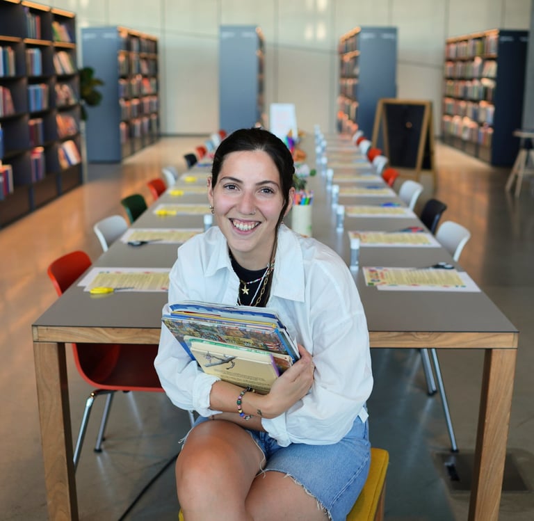 A smiling young woman holding books in a modern library with study tables and bookshelves.
