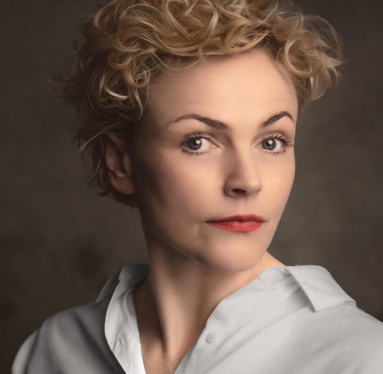 Professional studio headshot of a Maxine Peakewith short blonde curly hair and red lipstick.