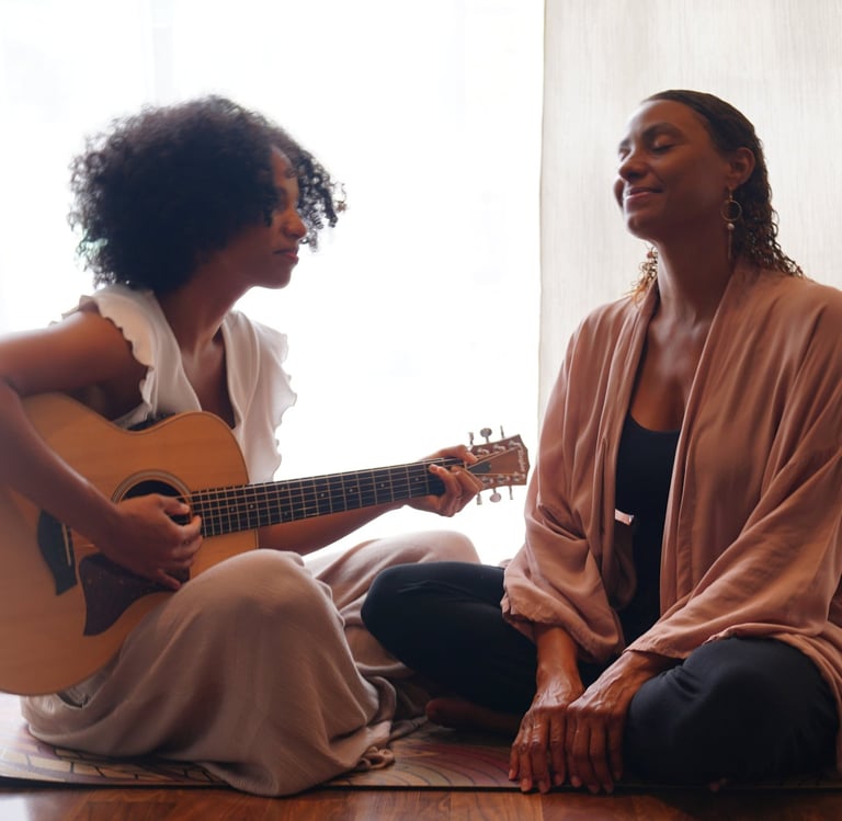 two women in front of a bright window sitting on a carpet playing a guitar.