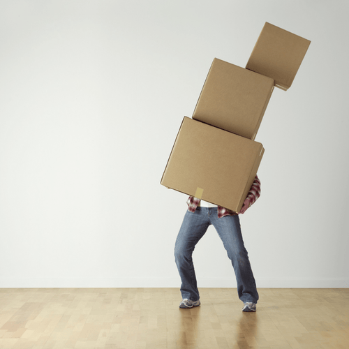 A man balancing a precarious stack of cardboard moving boxes in an empty room with hardwood floors.