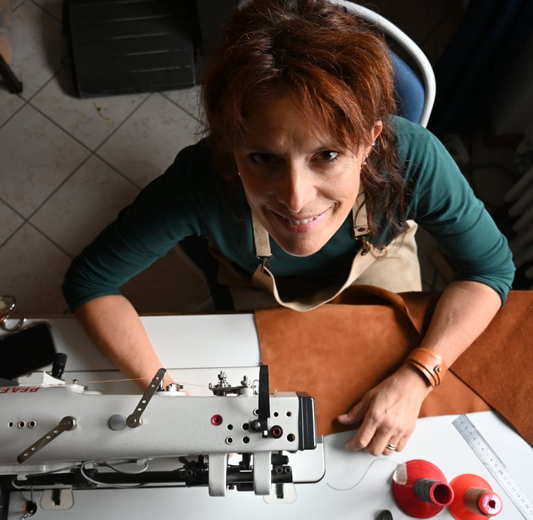 Female leather artisan smiling at her industrial sewing machine while crafting a handmade leather bag.