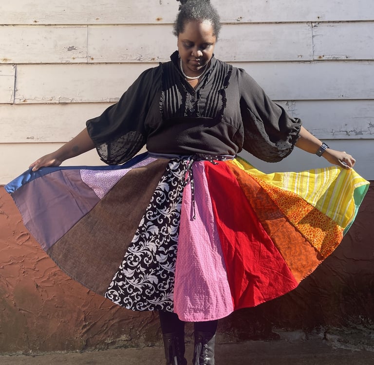 a black woman wearing a black shirt and rainbow circle skirt standing against a wall