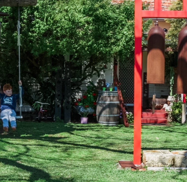 A young boy on a tree swing in a green garden with large hanging chimes and a red frame.
