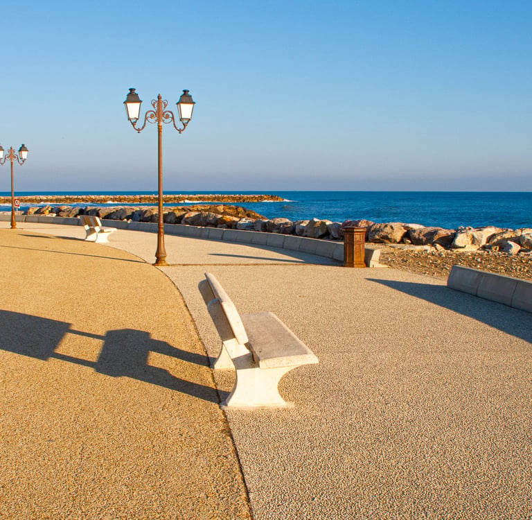 Empty stone benches and vintage street lamps along a paved seaside promenade with blue ocean views.