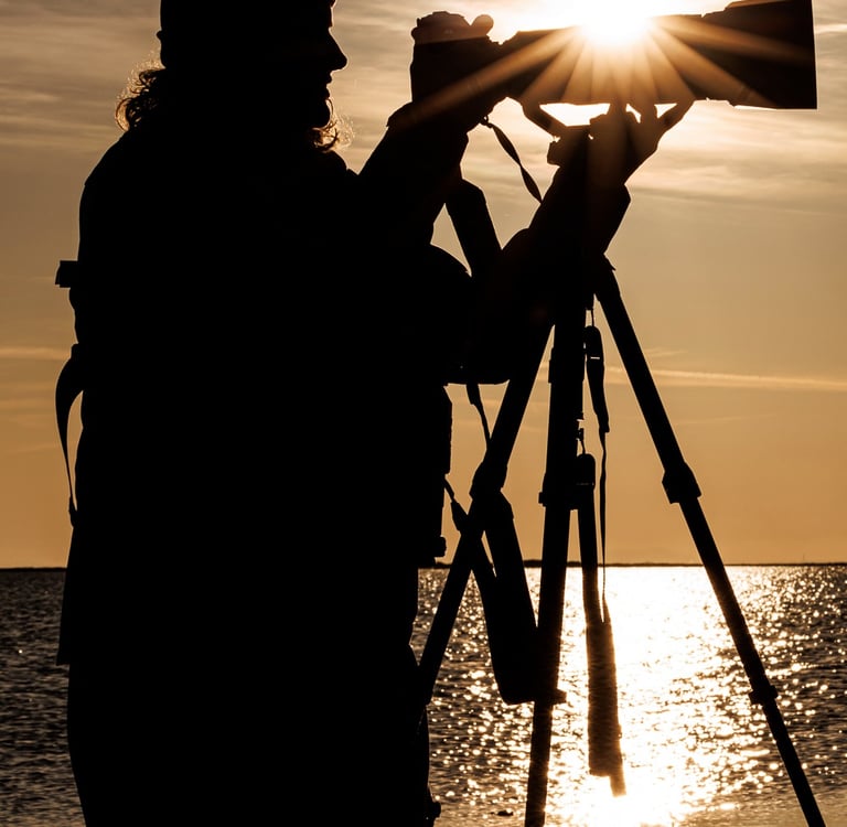 Silhouette of a photographer using a DSLR camera and tripod to capture a golden sunset over the ocean.