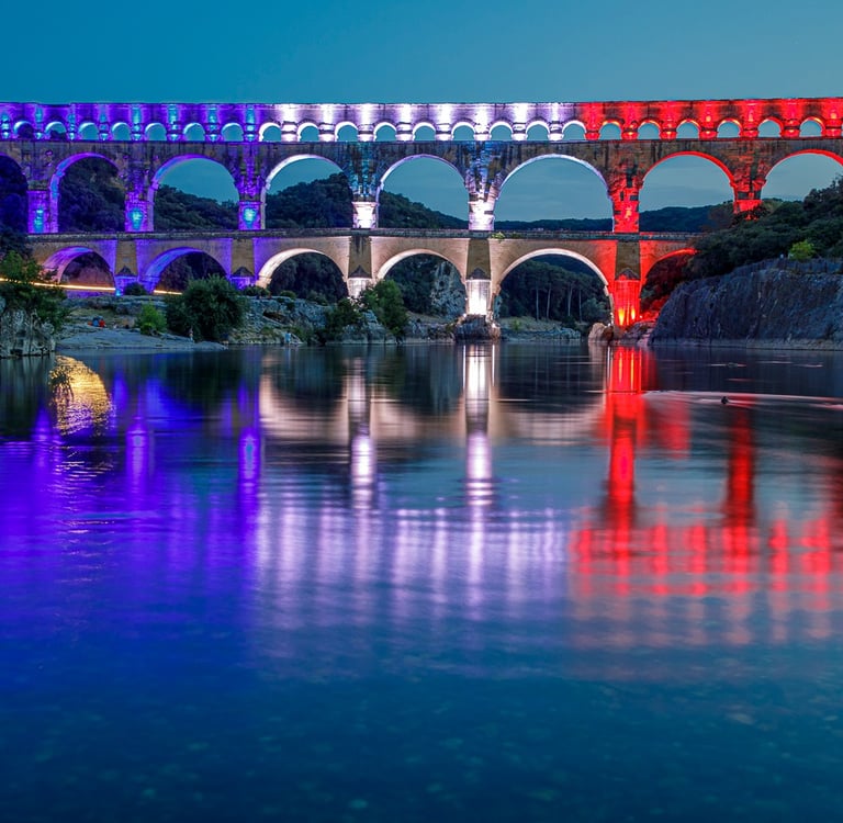 The Pont du Gard Roman aqueduct bridge illuminated in red, white, and blue lights reflecting on the Gardon River at night.