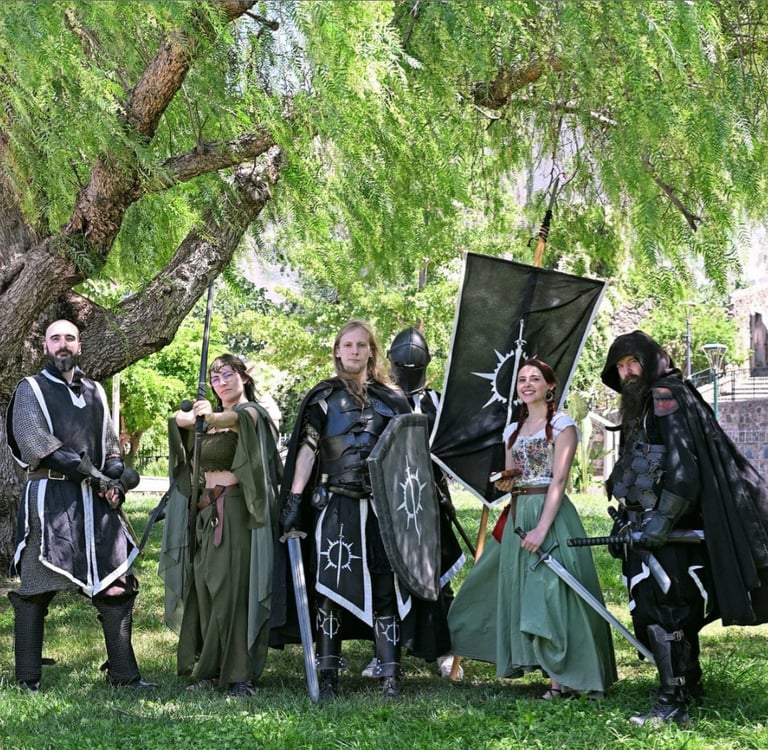 A group of medieval fantasy cosplayers in armor and robes holding swords and a banner under a large tree.