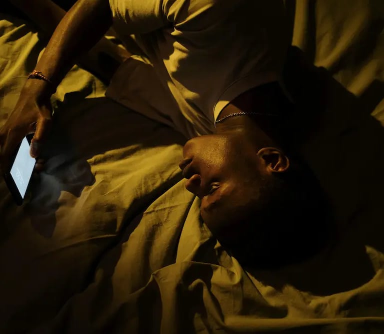 A man lying in bed at night using a glowing smartphone with dim, warm lighting.