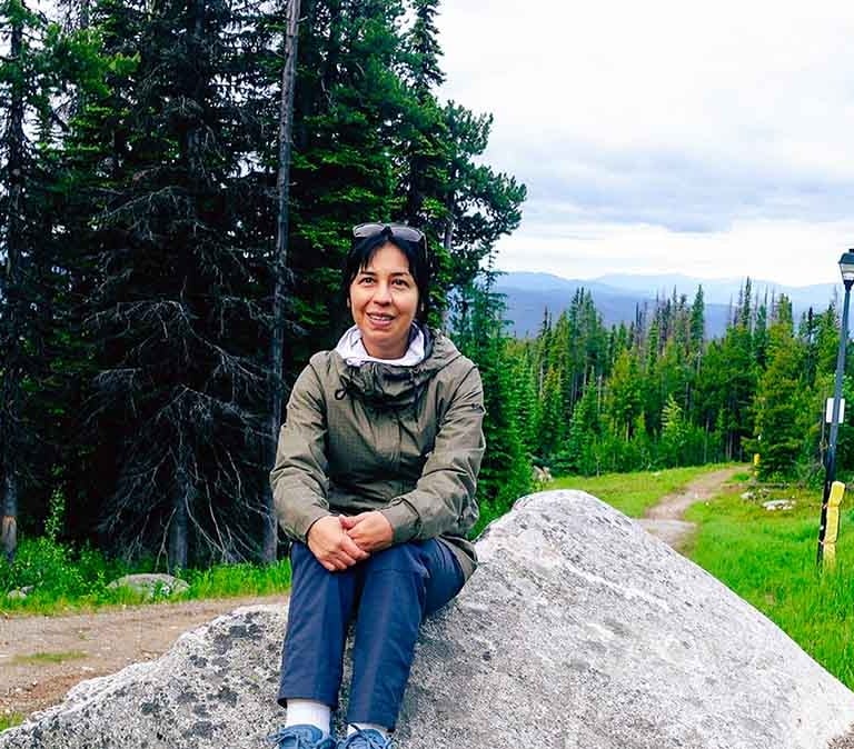 Woman sitting on a large rock beside a hiking trail surrounded by evergreen trees in Big White, Canada.