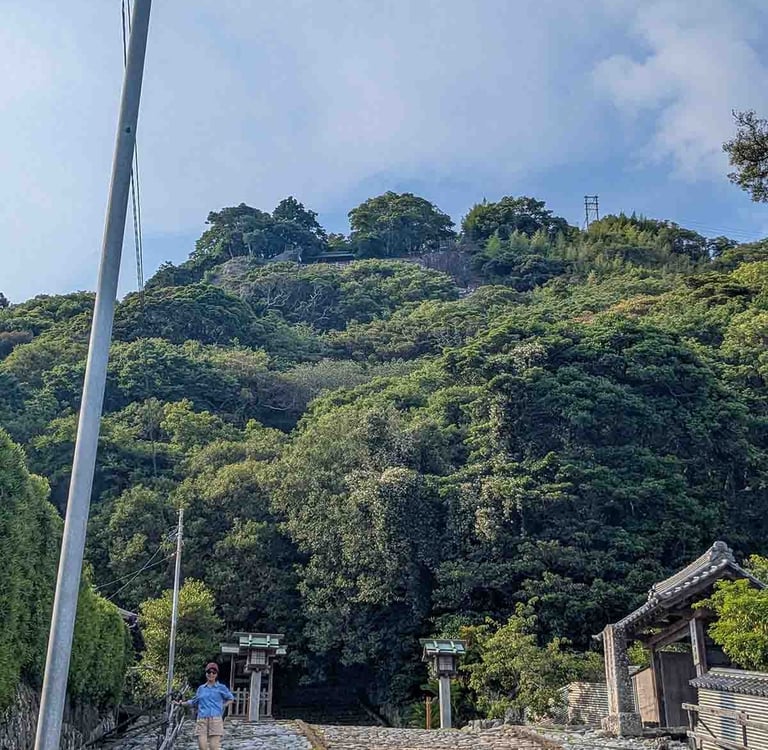Woman walking near a temple entrance beneath a lush green hillside in rural Shizuoka, Japan.