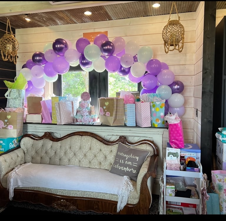 Purple and white balloon arch over baby shower gift table and vintage tufted sofa.