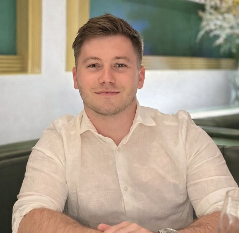 Harrison, smiling, wearing a white linen shirt and silver watch sitting at a restaurant table.
