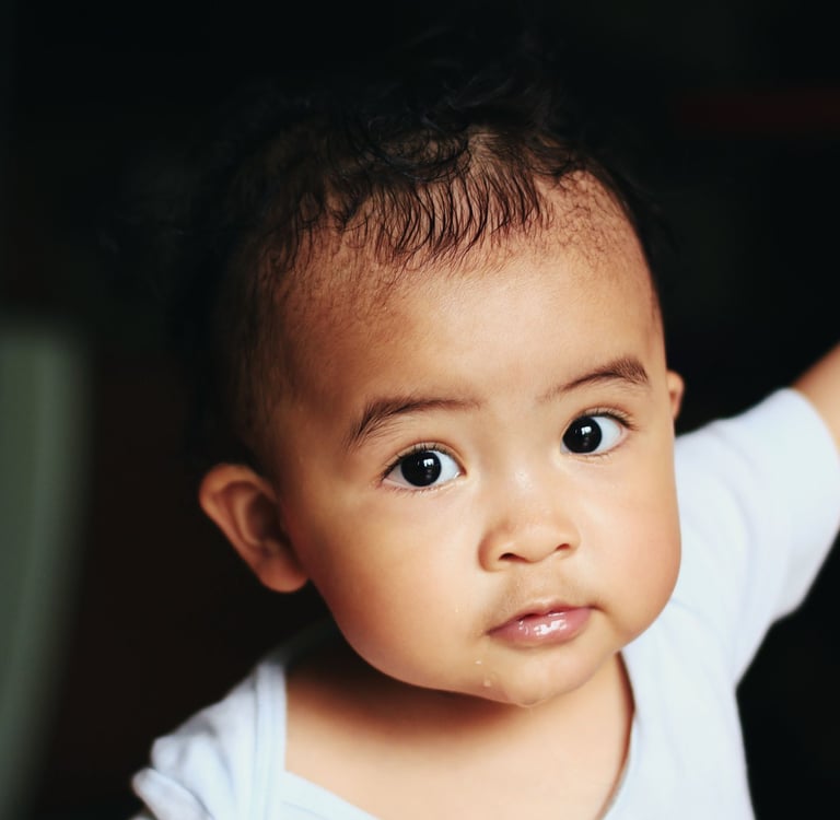 A close-up portrait of a cute baby with dark curly hair and large expressive eyes wearing a white shirt.