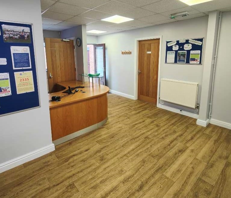 A reception with curved wood desk, wooden flooring/door, and light-grey walls.
