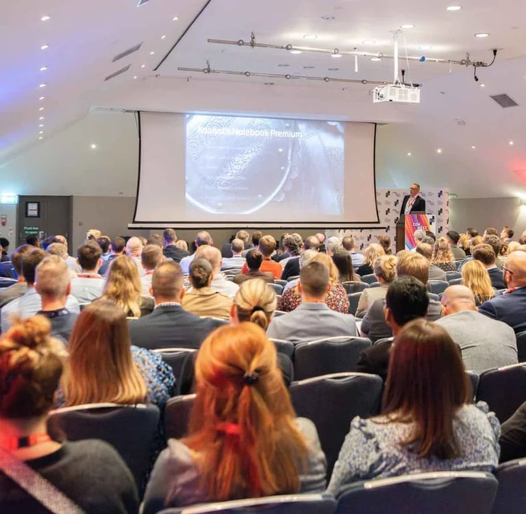 A speaker presents at a business conference with an audience seated in a large auditorium.