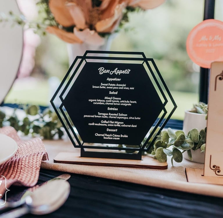 Elegant black hexagonal wedding menu card on a rustic table with greenery and place settings.