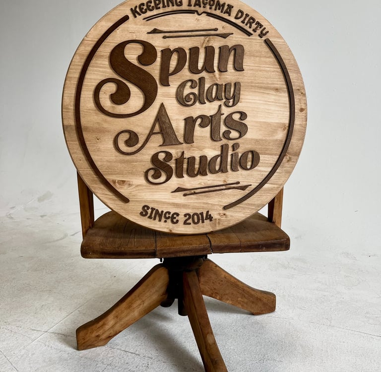 Custom wood round sign for Spun Clay Arts Studio resting on a vintage wooden pottery stool.