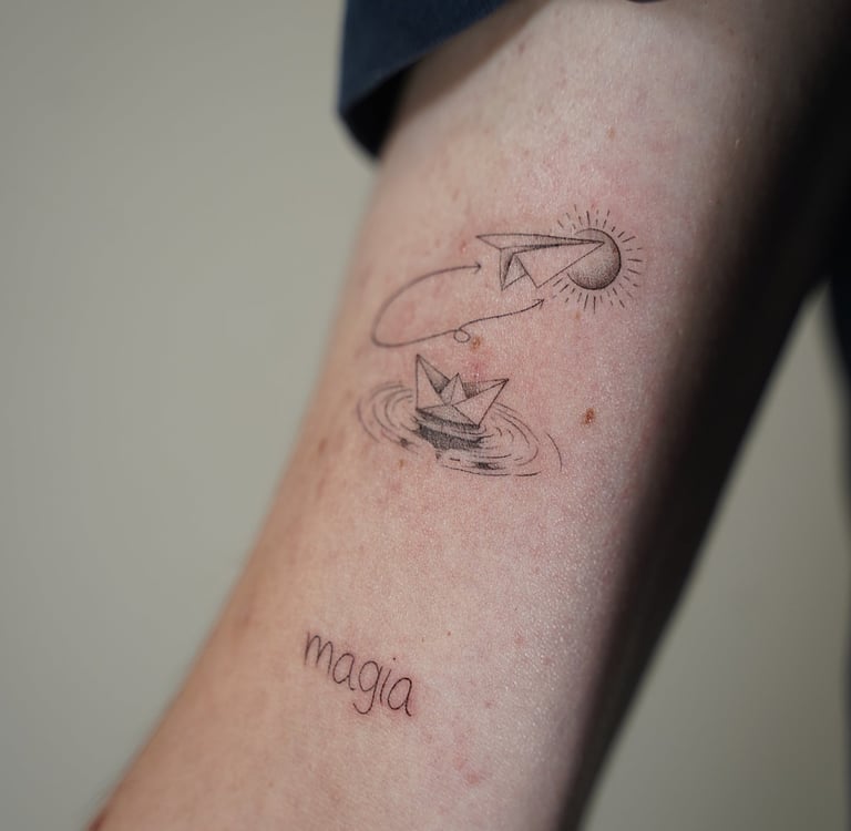 Tatuaje fineline en Madrid de barco de papel y avión de origami con luna y la palabra magia